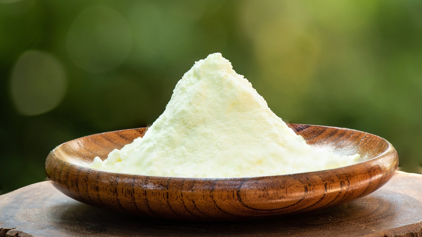 A shallow wooden bowl holds a mound of soft, light green powder, its fine texture subtly reflecting light, set against a blurred natural green background.